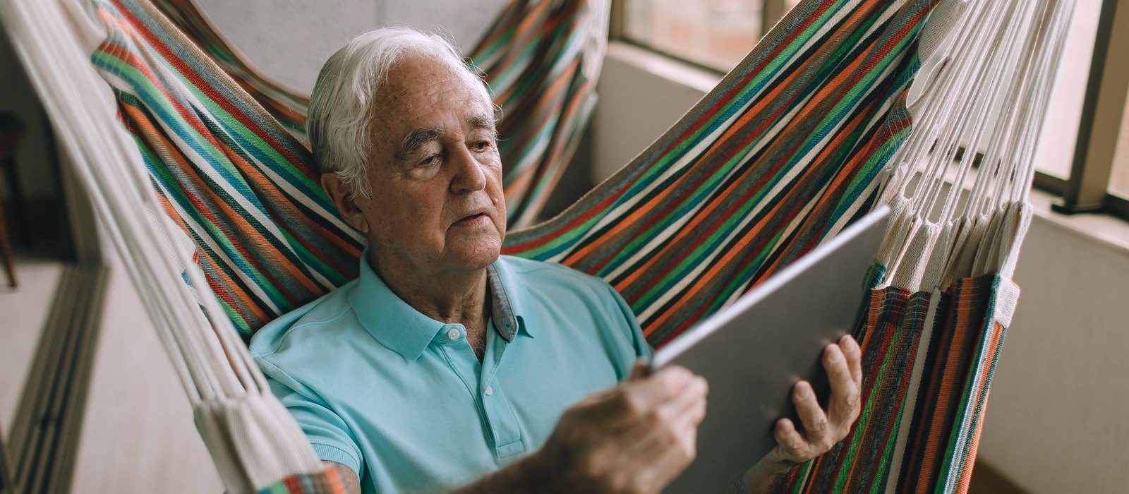 A white-haired man in a light blue polo shirt sits in a brightly striped hammock chair on a balcony, reading a tablet he holds in both hands