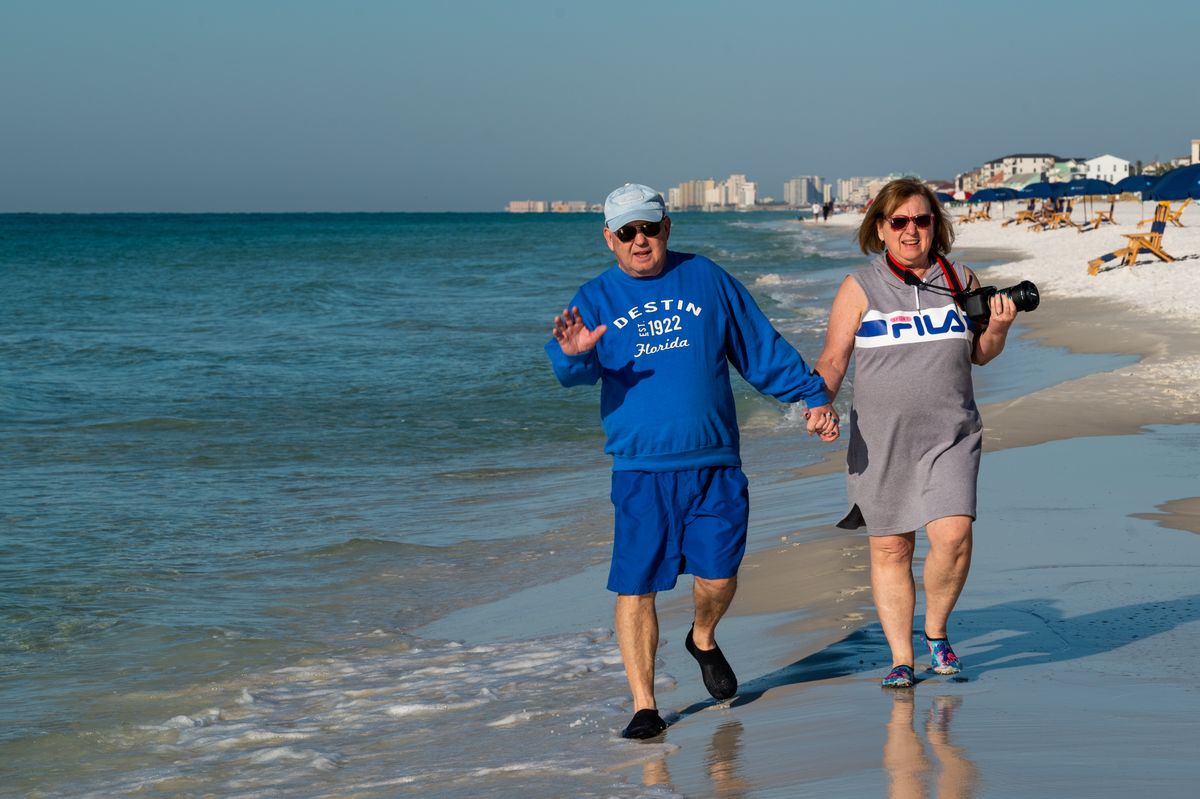 A retired couple hold hands walking along the surf on a wide white-sand Florida beach, the woman carrying a camera