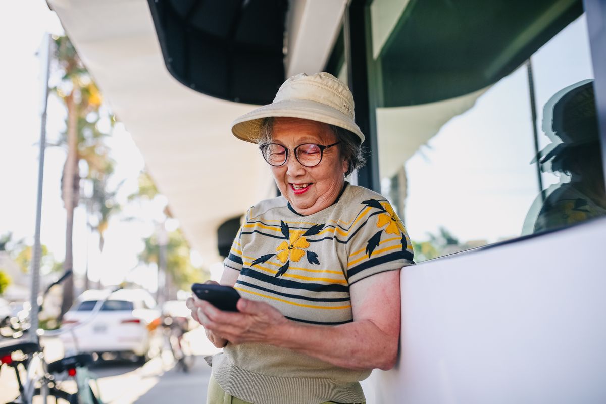 A smiling older woman in a sun hat and glasses reads her phone on a sunny sidewalk, palm trees and parked cars behind her
