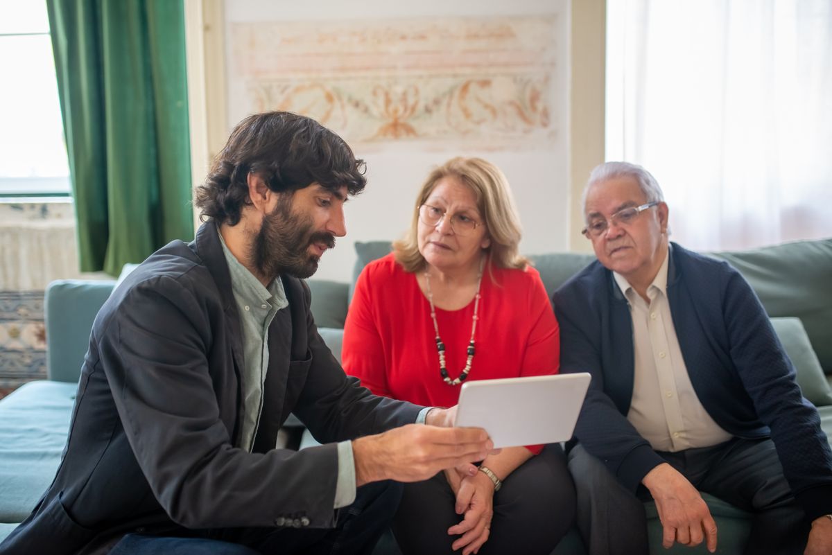 A younger man in a dark jacket shows a tablet to an older couple sitting together on a sofa in a sunlit living room