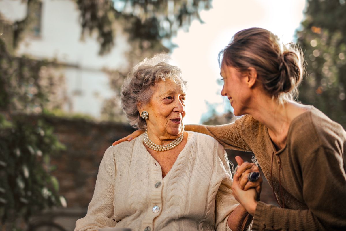 An adult daughter rests her arm around her elderly mother's shoulders in a sunlit garden while the two talk