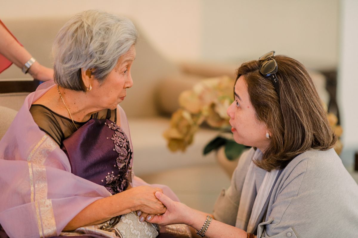 A grey-haired woman in a lilac shawl and her adult daughter sit close together in a living room, holding hands as they talk