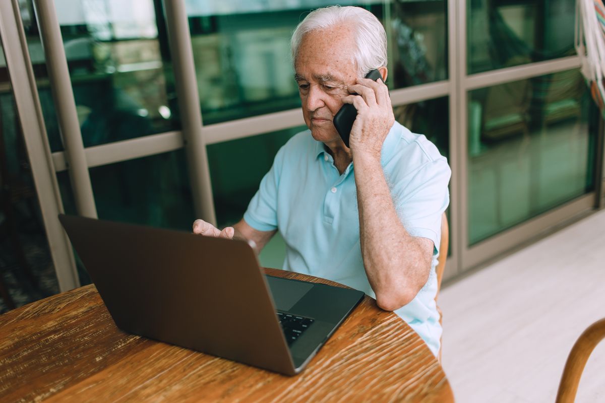 A white-haired man in a light blue polo shirt sits at a wooden table with an open laptop, talking on his phone by a glass patio door