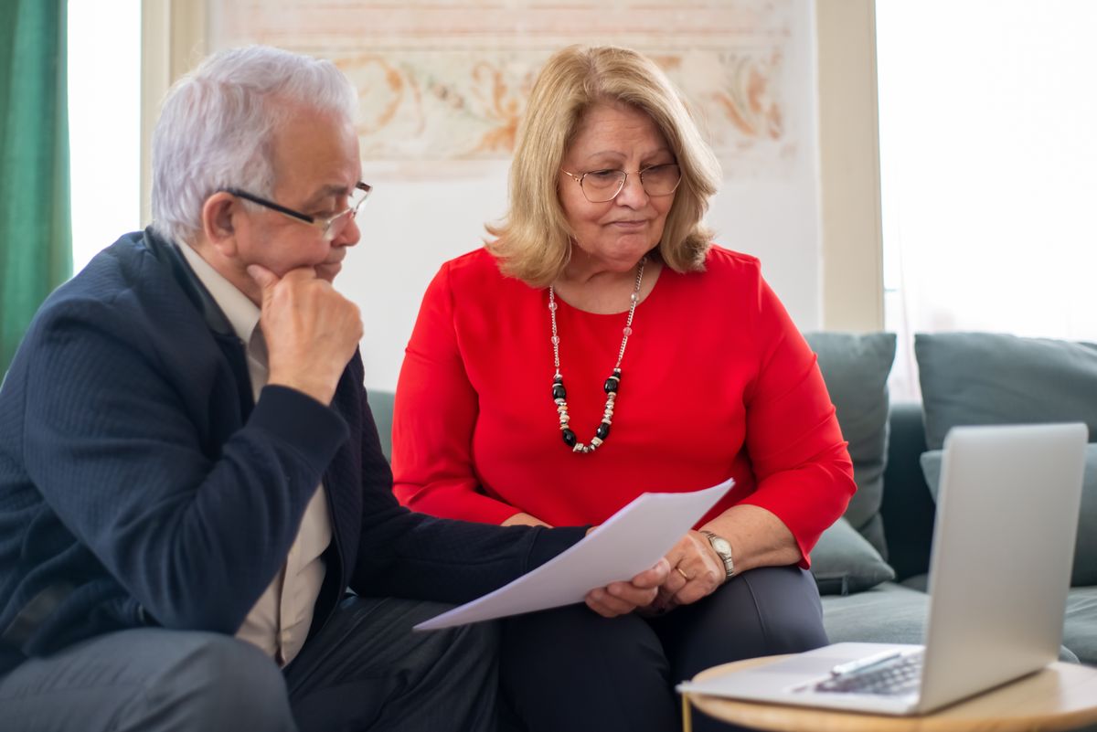 A retirement-age couple sit together on a sofa reading a document, with a laptop and calculator on the side table beside them