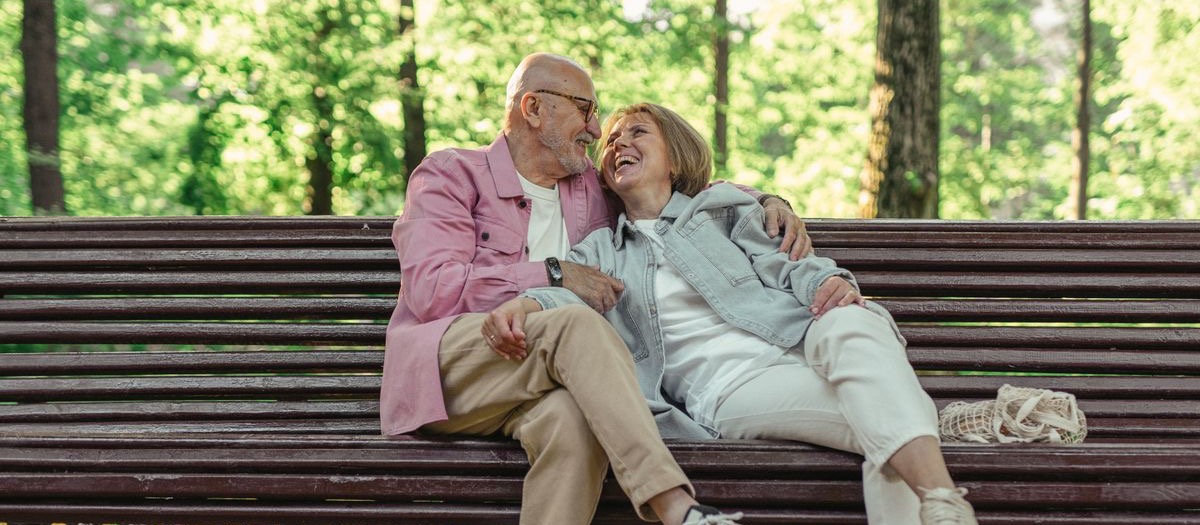 A retired couple laugh together on a wooden park bench under summer trees, the man in a pink shirt with his arm around the woman's shoulders