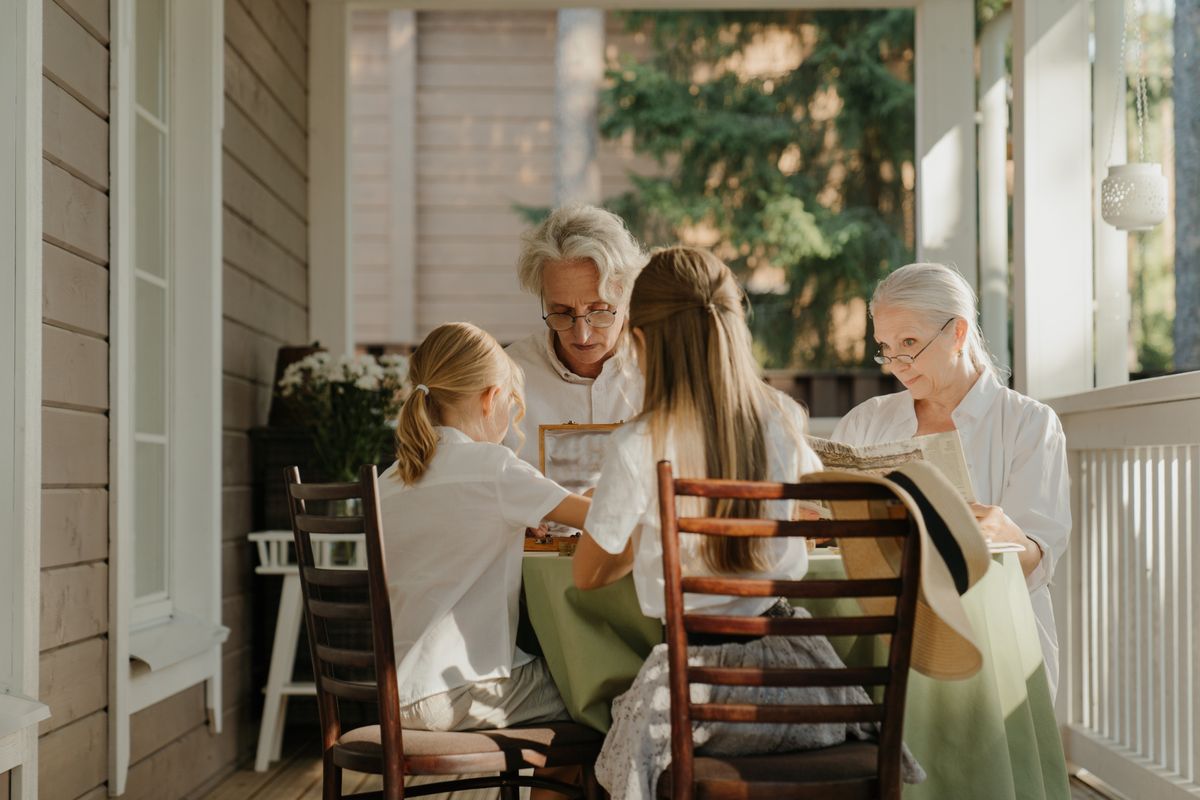 Two grandparents and two grandchildren gathered around a table on a shaded wooden porch on a sunny afternoon