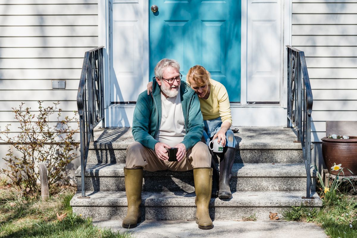 An older couple in rain boots sit close together on their front steps with mugs of coffee on a sunny morning