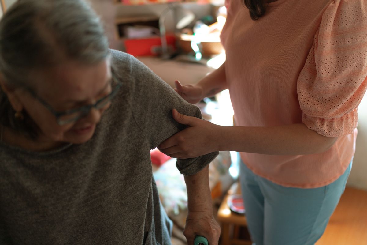 A woman in a peach blouse steadies an older woman's arm as she stands with a cane in a sunlit living room