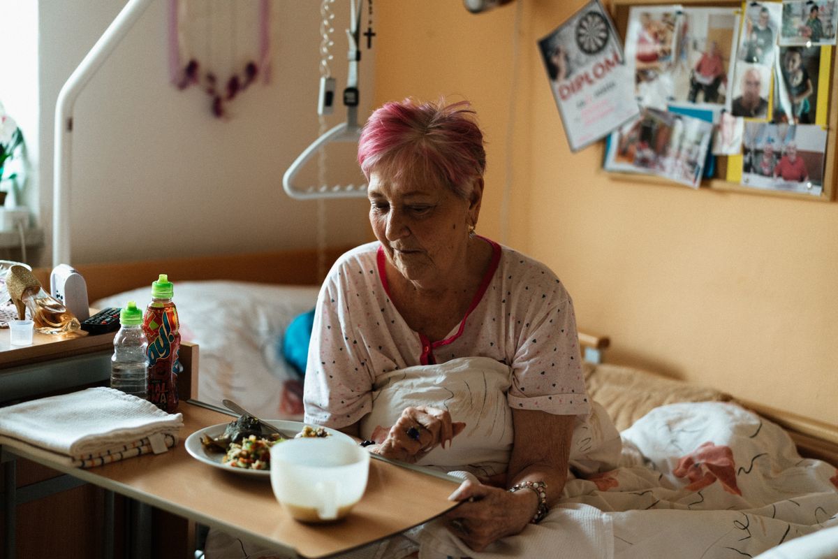 An older woman sits up in bed eating a meal in her room at a care facility, family photographs pinned to the board above her