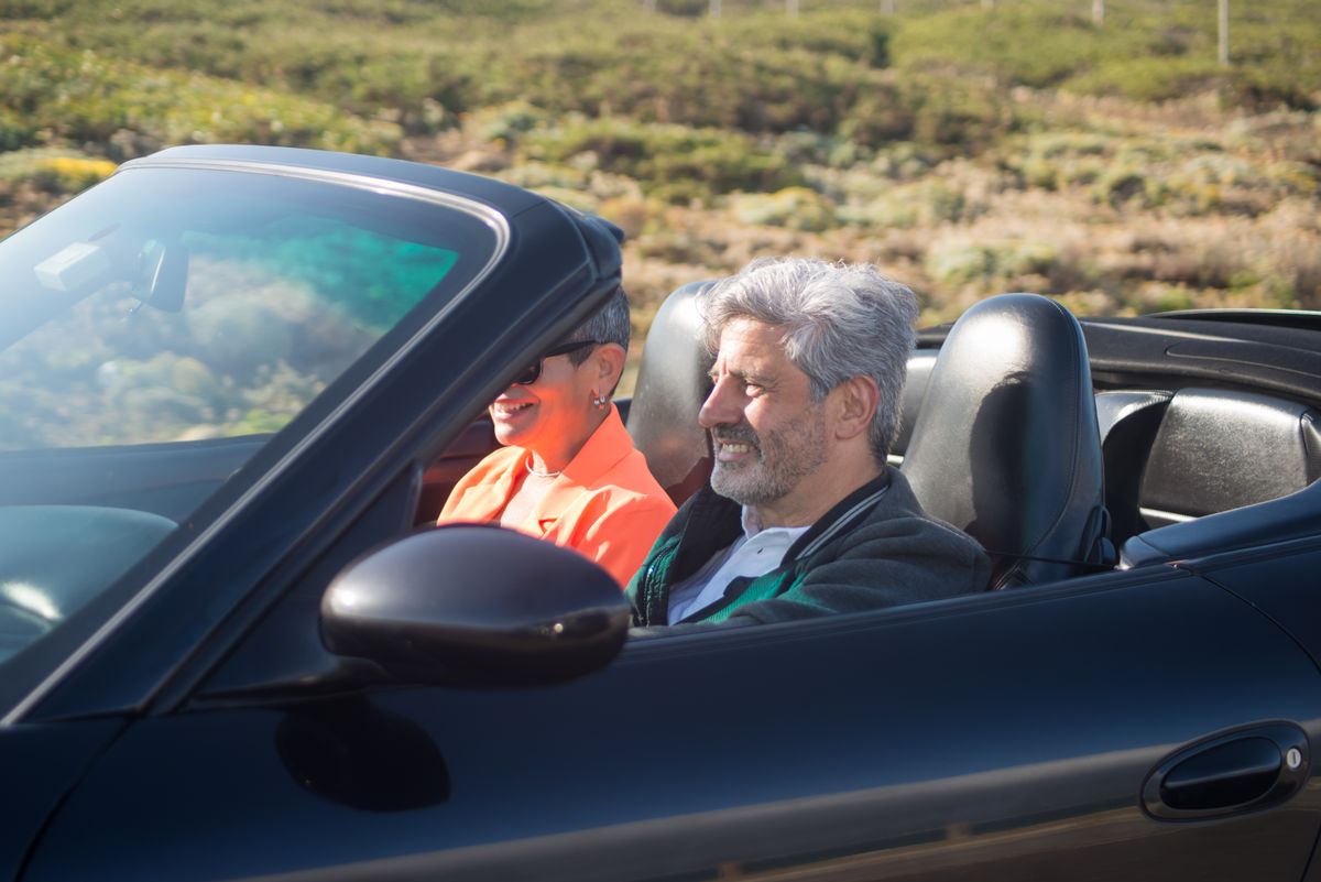 A grey-haired couple drive a convertible along a coastal road on a bright day, the passenger in an orange jacket and sunglasses
