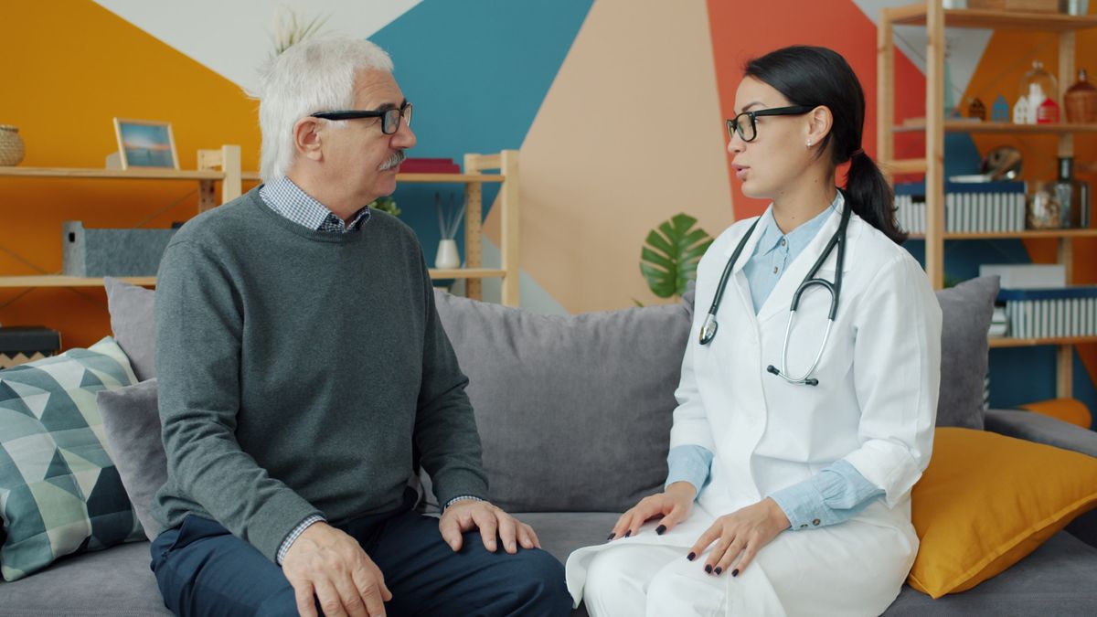 A white-haired man in glasses and a grey sweater sits on a sofa talking with a doctor in a white coat with a stethoscope around her neck