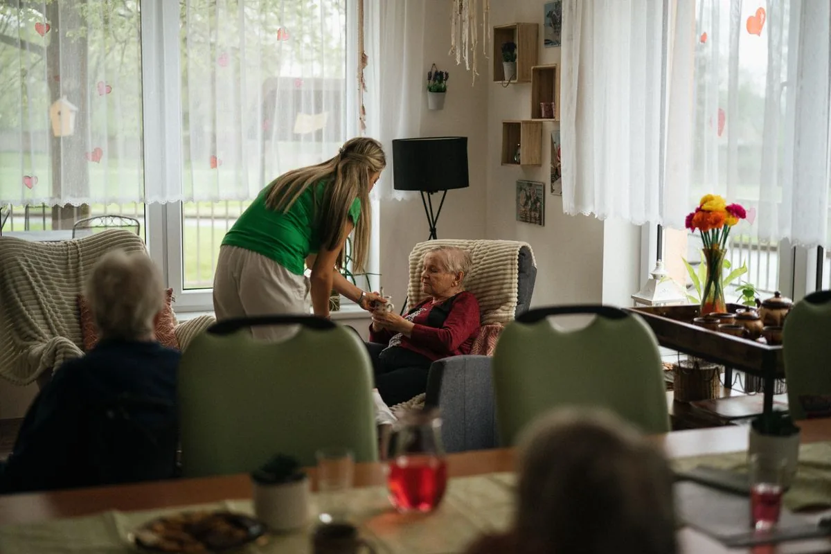 A caregiver brings tea to a seated older woman in the bright common room of an assisted living community