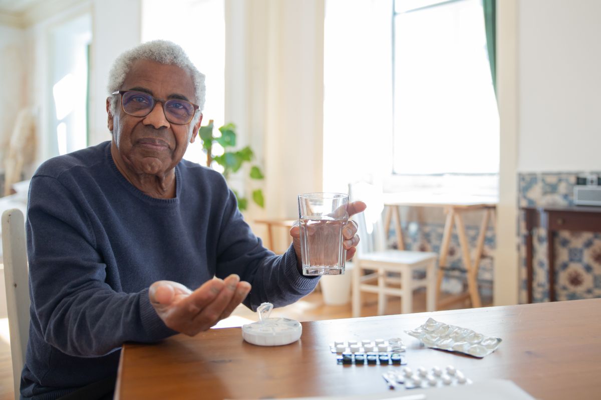 An older man in a navy sweater sits at a sunlit kitchen table holding a glass of water, with a pill organizer and blister packs in front of him