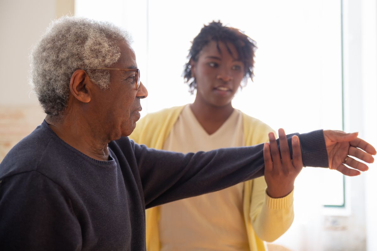 An older man in glasses extends his arm through a stretch while a therapist steadies his wrist in a sunlit room