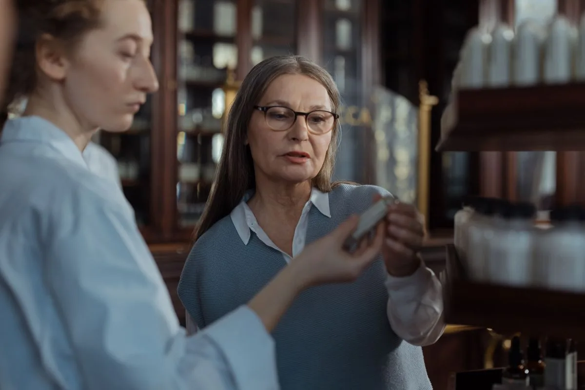 A pharmacist hands a medication box across the counter to an older woman reading the label