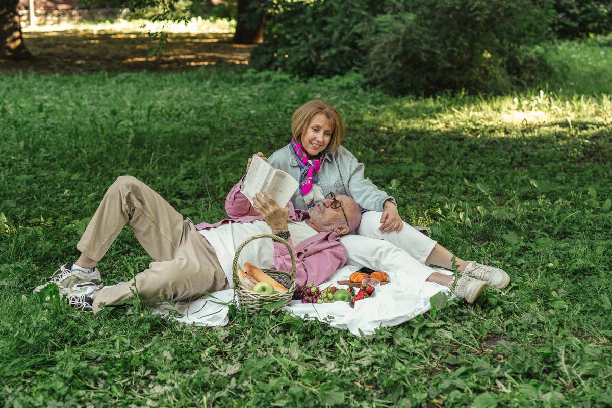 A grey-haired couple on a picnic blanket in a shaded park, the man lying back reading a paperback with his head in his wife's lap, a basket of bread and fruit beside them