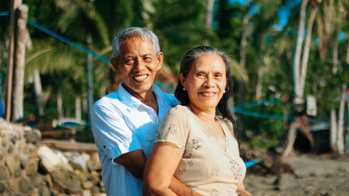 A smiling retired couple stand close together in the sunshine with palm trees behind them, the man in a light blue shirt and the woman in a floral top