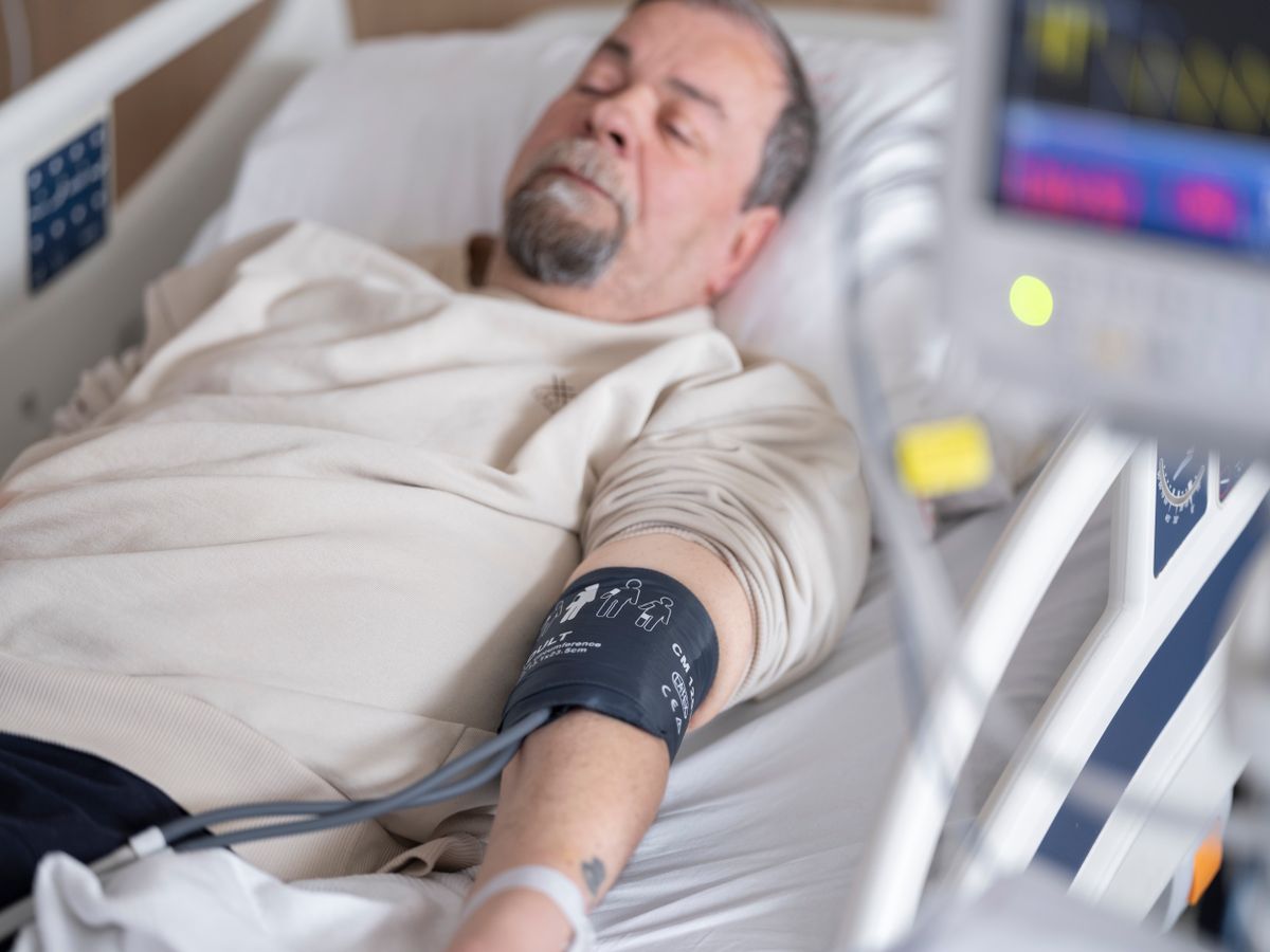 An older man with a grey beard resting in a hospital bed with a blood pressure cuff on his arm and a monitor beside him