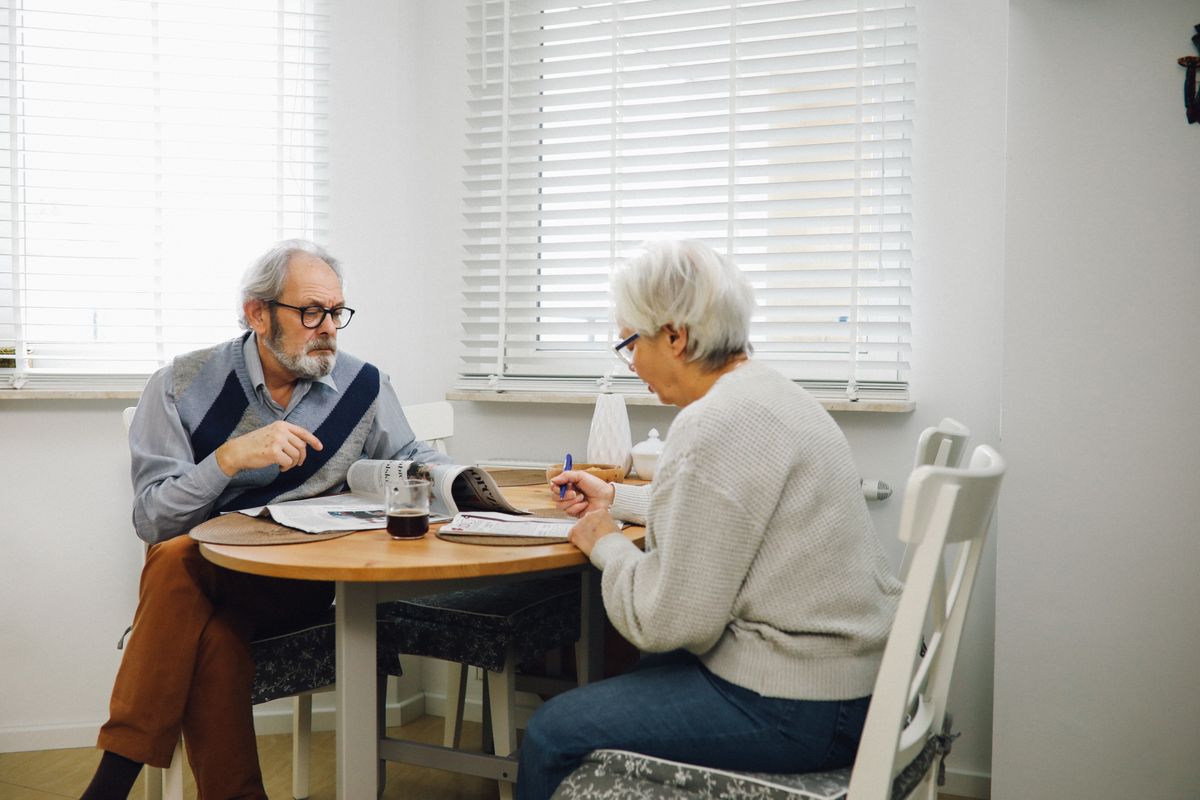 An older couple sit across from each other at a round kitchen table beside a bright window, the man reading a newspaper and the woman writing with a blue pen