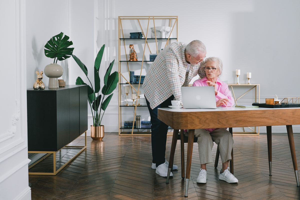 An older couple study a laptop together at a wooden dining table at home, the woman seated in a pink shirt and the man leaning in over her shoulder.