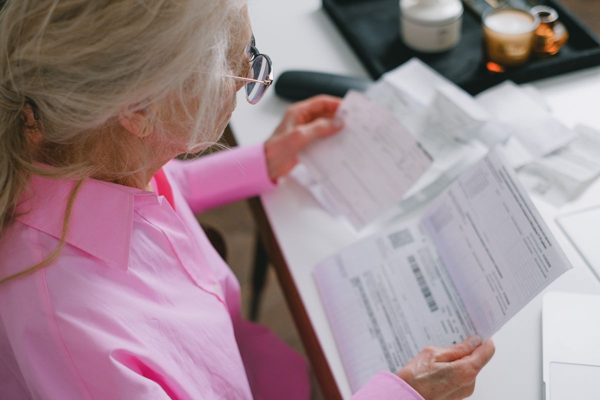 An older woman in a pink shirt and glasses sits at a white table looking over a handful of itemised medical bills