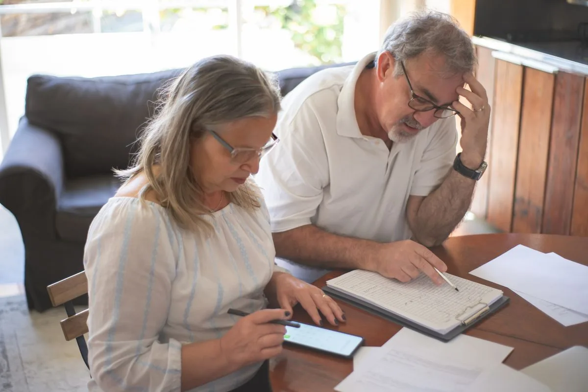 An older couple work through insurance paperwork together at their dining table, one holding a pen over a clipboard