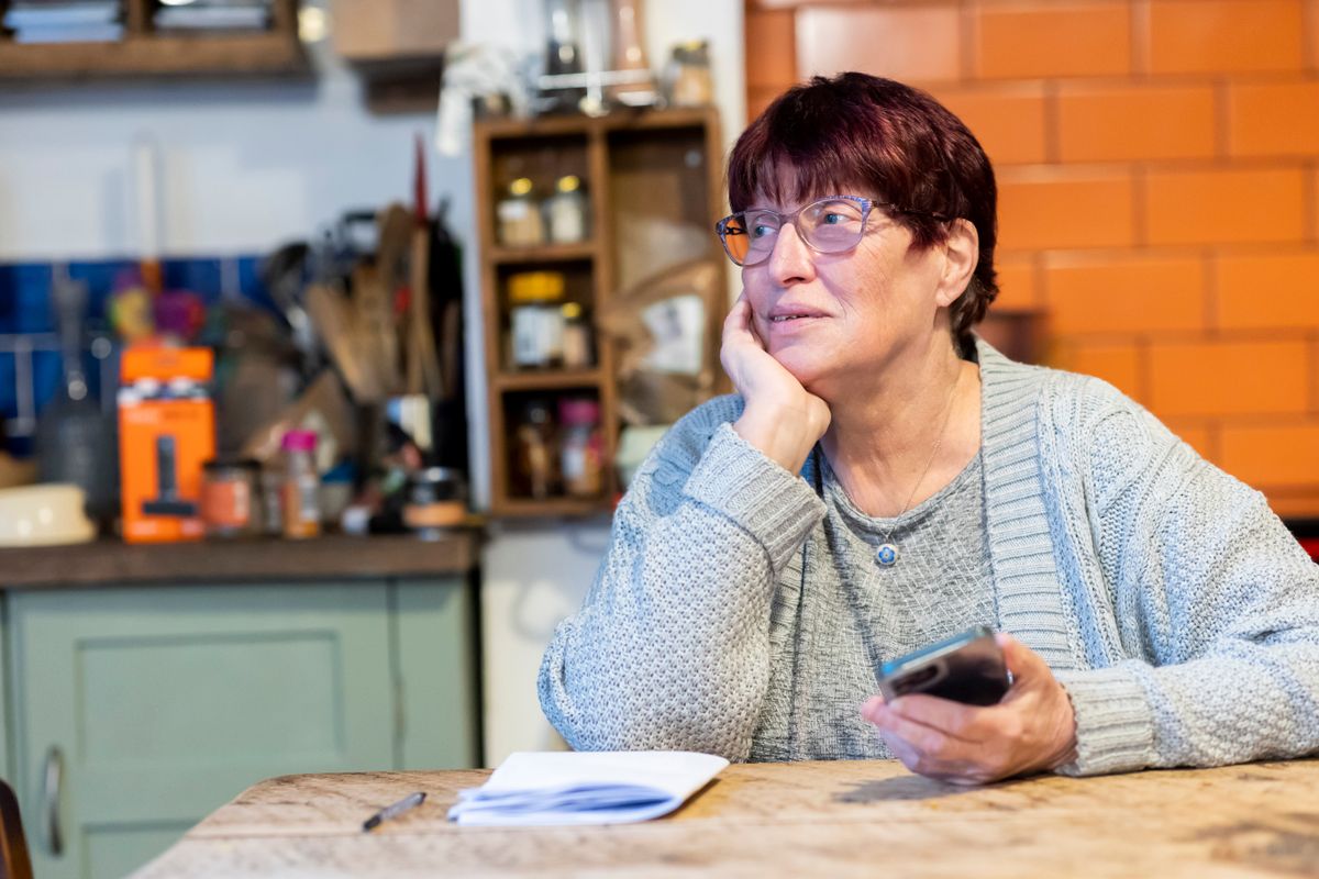 An older woman in a blue cardigan sits at her kitchen table with a stack of paperwork and a pen, holding her phone and looking away in thought