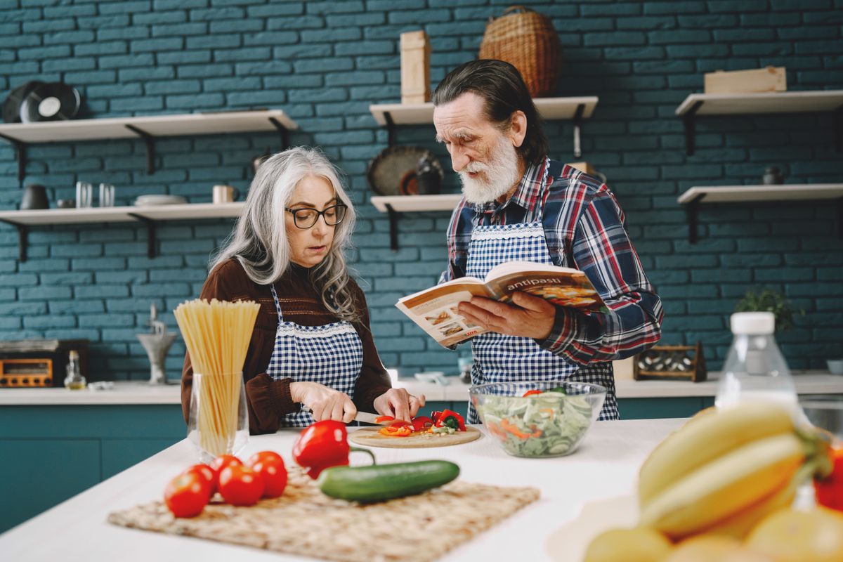 A grey-haired couple in checked aprons cook together at a kitchen island, the woman slicing peppers while the man reads a recipe book