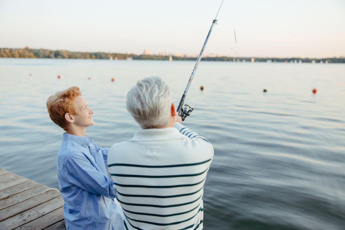 A white-haired grandfather in a striped shirt casts a fishing rod from a wooden dock at sunset while his teenage grandson looks on