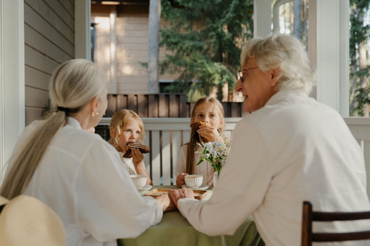 Grandparents share coffee and pastries with two young granddaughters at a table on a shaded porch