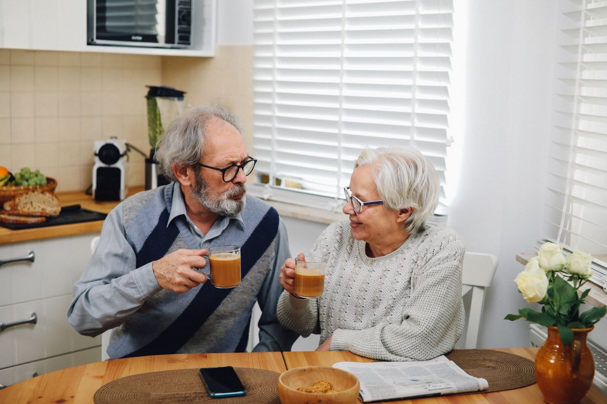 A grey-haired couple in glasses and knitwear raise glass mugs of coffee to each other at a wooden kitchen table, a newspaper and a vase of white roses beside them
