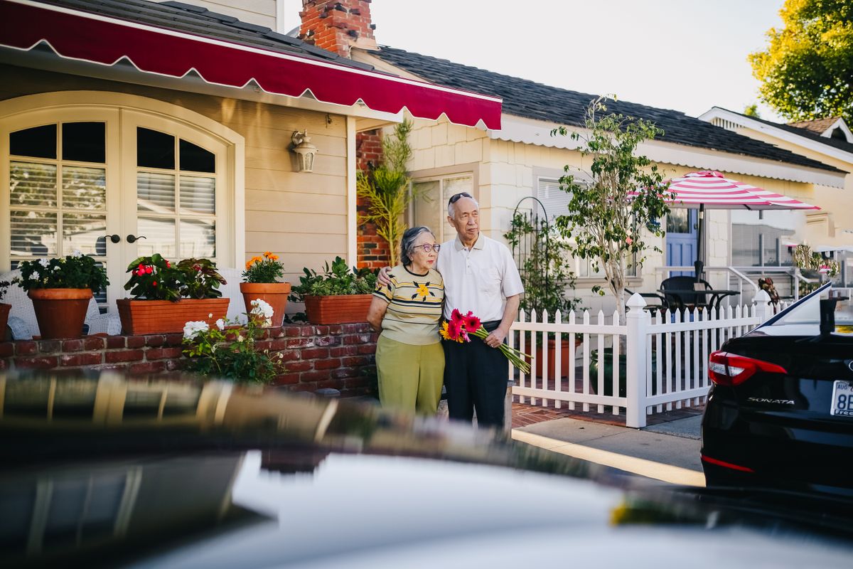 An older couple stand arm in arm outside their single-storey home beside a white picket fence and potted flowers, the man holding a bunch of red gerberas