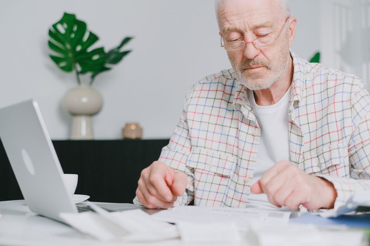 An older man in glasses and a checked shirt reads paperwork spread across a table beside an open laptop and a cup of coffee.