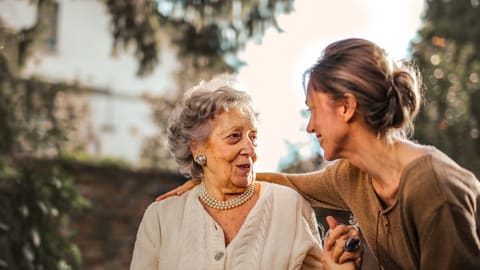 An adult daughter greets her mother in a sunlit garden, both of them laughing