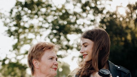 An older woman and a younger woman walk arm in arm outdoors, talking and smiling