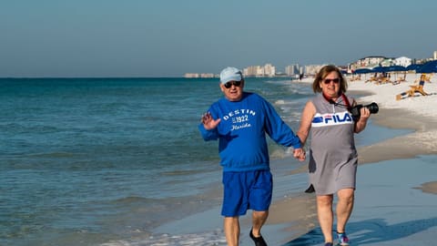 Two retirees in shorts and sun hats walk along the water's edge on a bright Destin, Florida beach