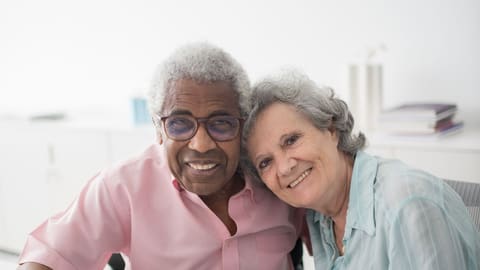 An older couple sit close together at a table, smiling, as one of them signs a form